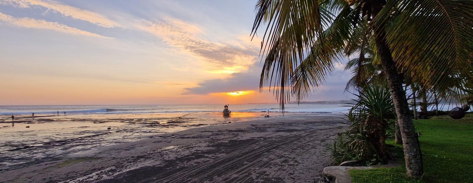 Zonsondergang boven de westkust van Bali, met donker zand, een palm en een jukung in de branding